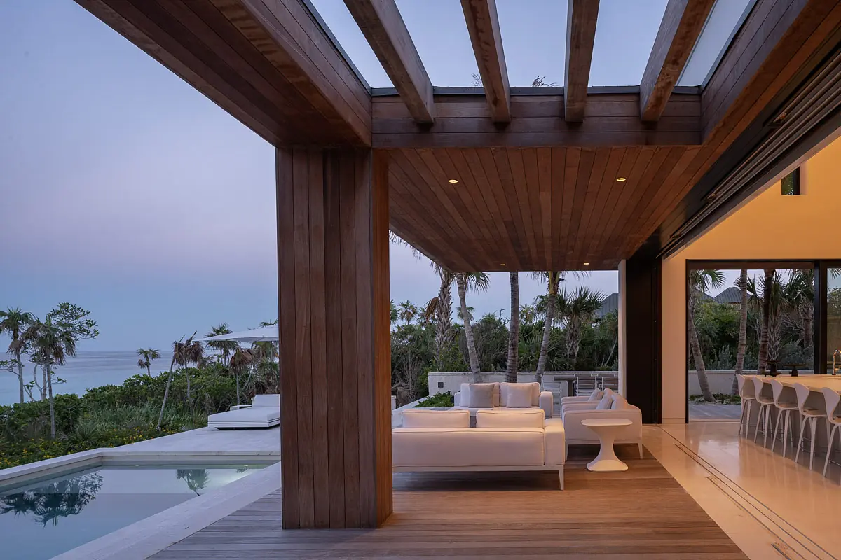 Deck with white sectional sofa, round side table, swimming pool, wooden beams, and tropical landscaping.