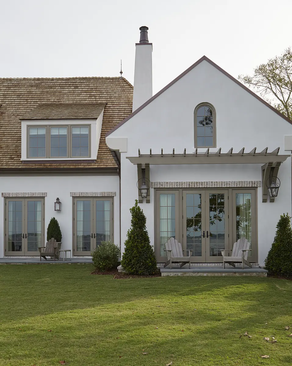 Exterior rear view of a house with white walls, glass doors, chimney, chairs, and shrubs on a grassy lawn.