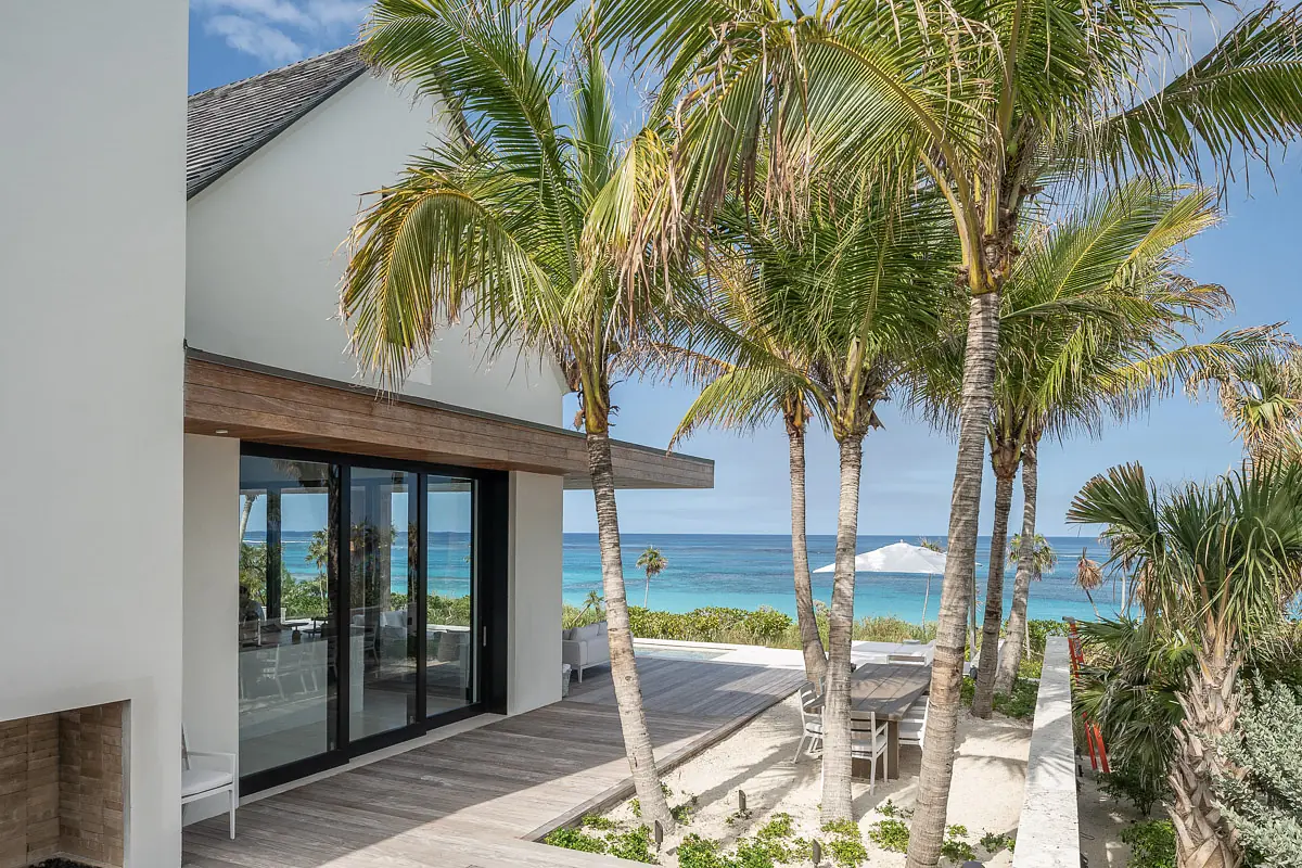 Beachfront exterior with palm trees, wooden deck, glass doors, and ocean view.