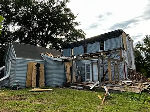 Partially demolished house with blue siding, exposed framing, tarp-covered roof, and debris in the yard.