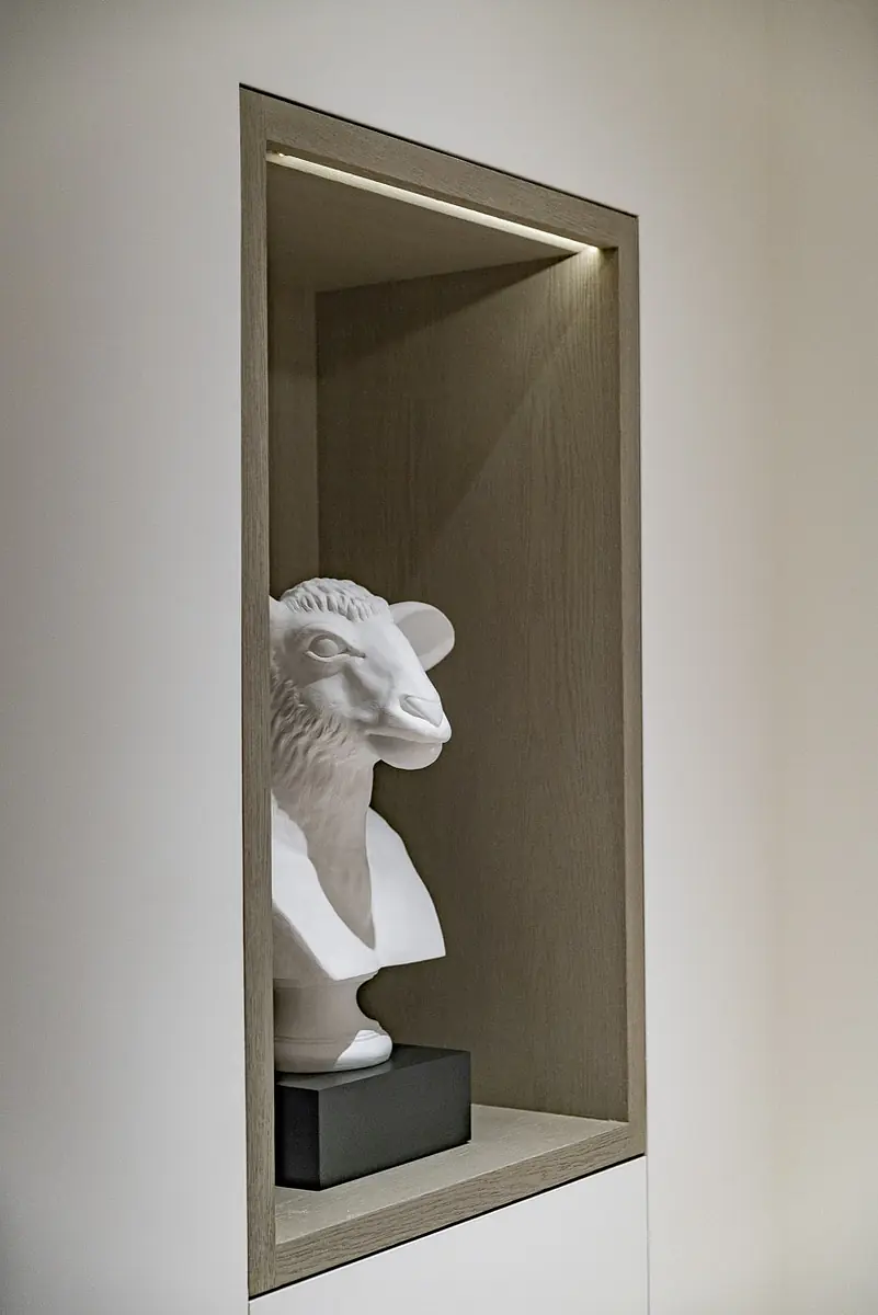 Niche with white sheep bust sculpture on a black base and lighted shelving.