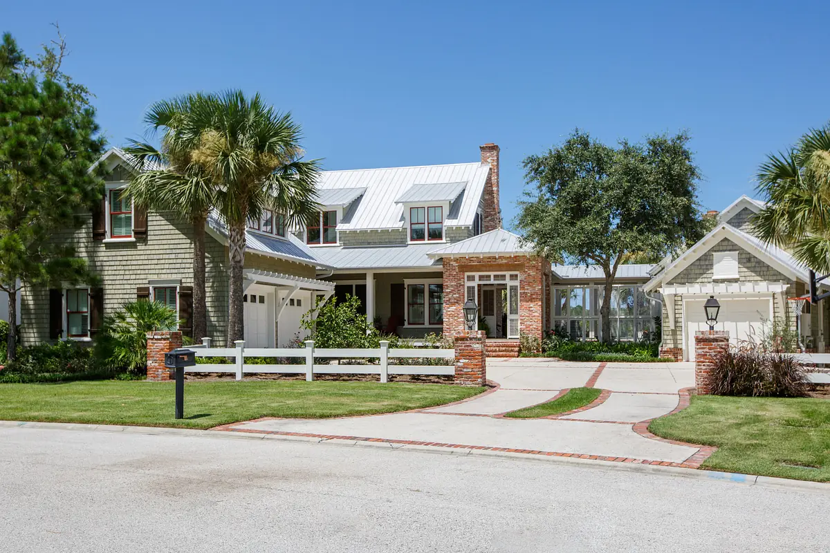 Exterior front of a house with paved driveway, grass lawn, palm trees, brick and wood siding, and metal roof.