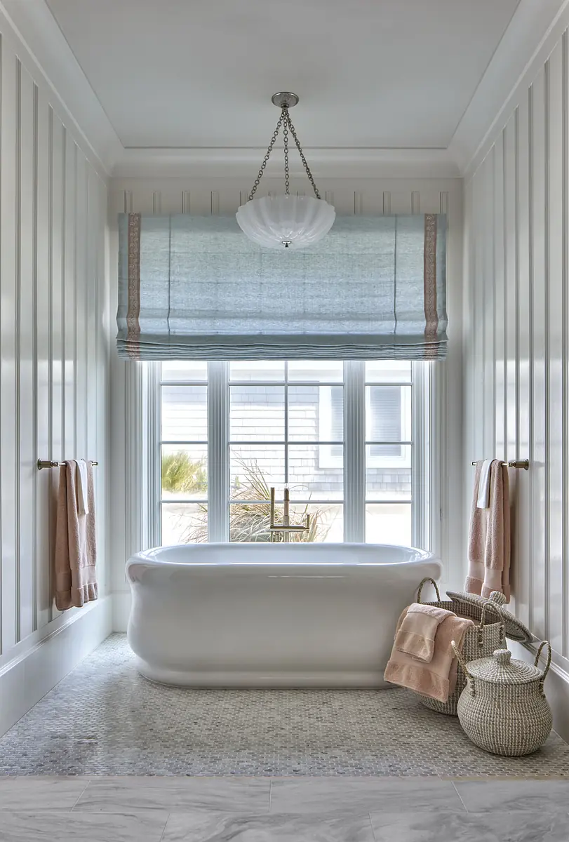 Bathroom with freestanding white bathtub, stone floor, blue window treatment, and pastel towels.