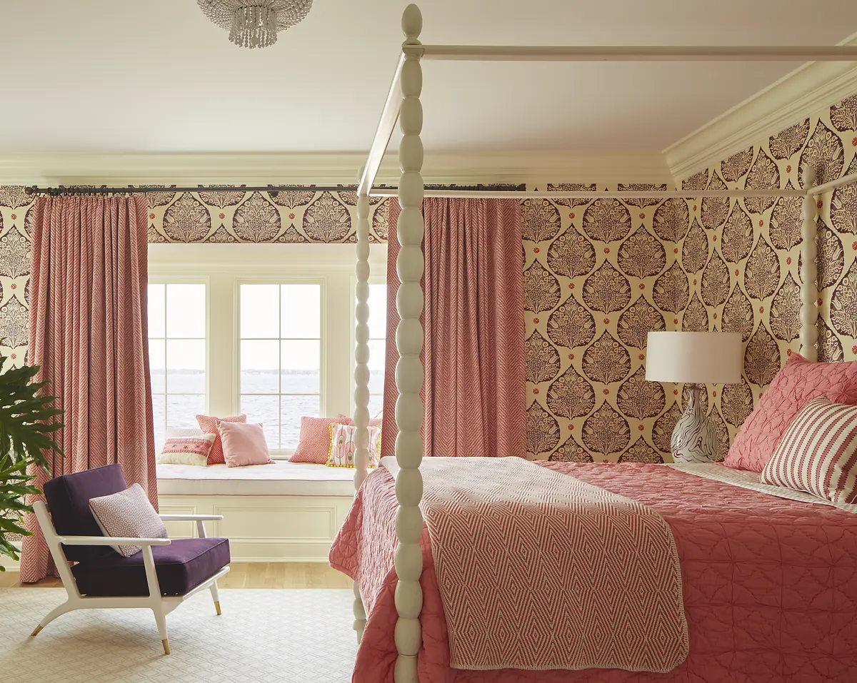 Bedroom with four-poster bed, pink quilt, purple armchair, patterned wallpaper, and window seat.
