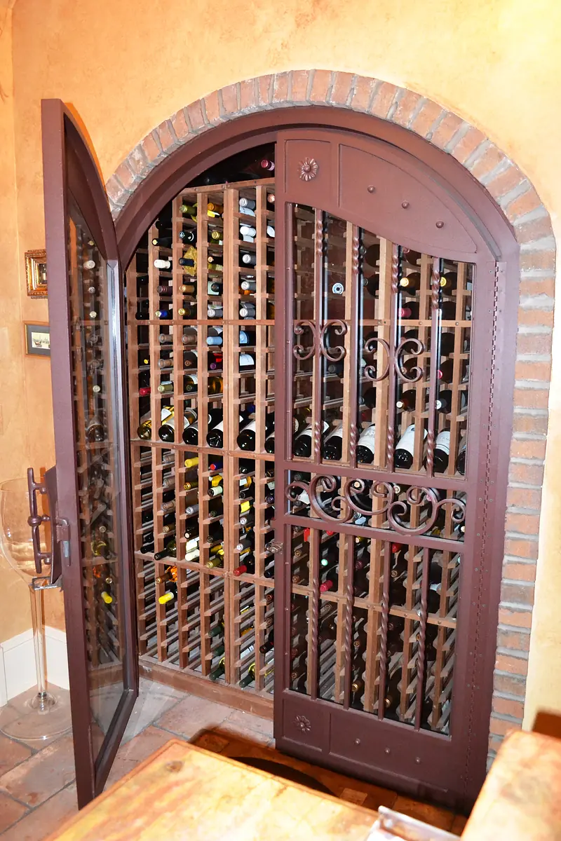 Wine cellar with double door, metal grills, wine racks, and terracotta tiled floor.