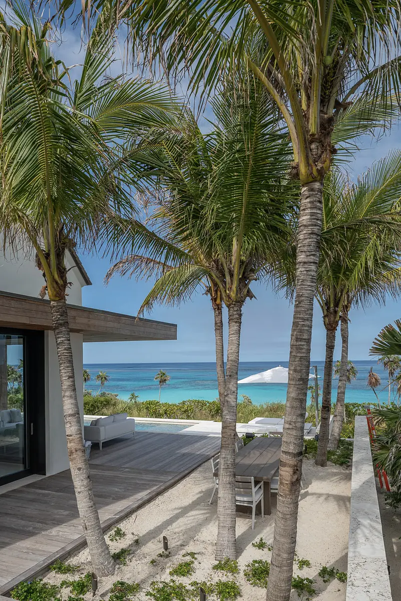 Patio with wooden flooring, dining table, white lounge chairs, and surrounding palm trees with ocean view