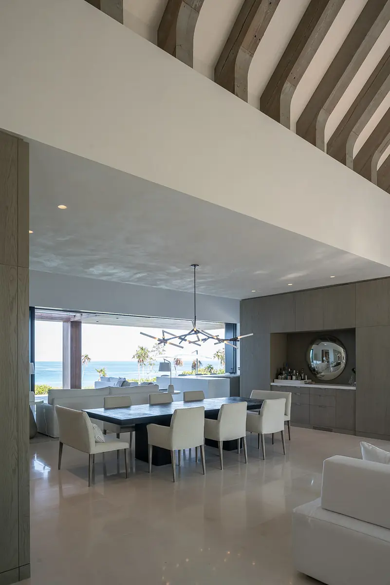 Dining room with large table, white chairs, ocean view, circular mirror, and exposed beam ceiling