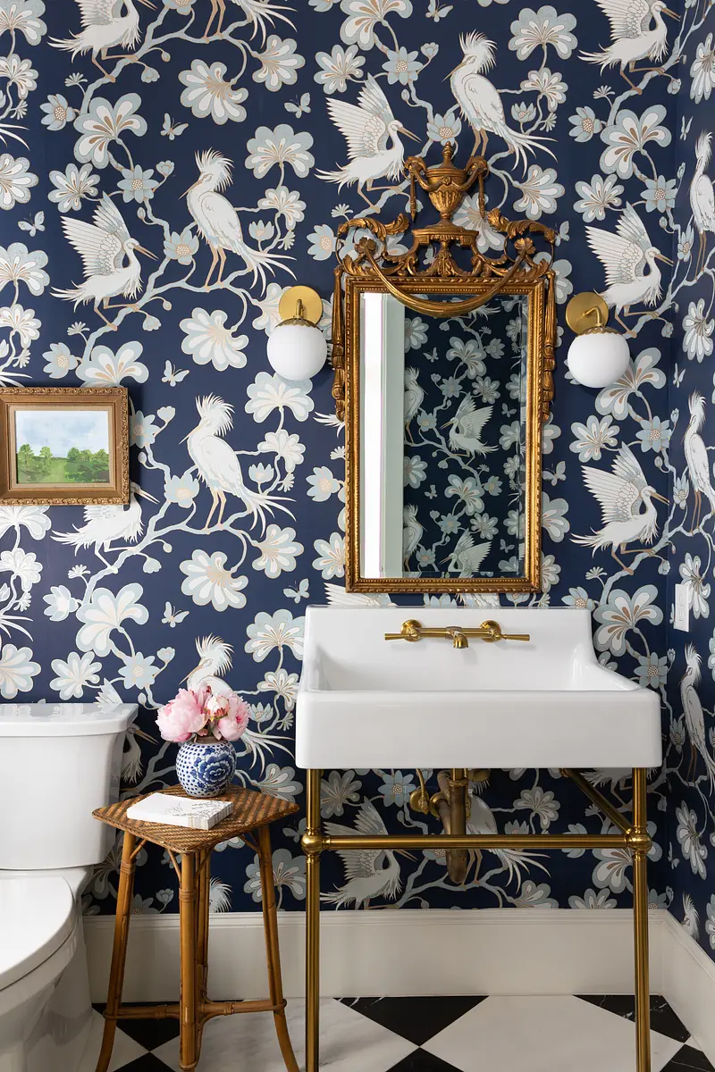 Bathroom with white pedestal sink, gold-framed mirror, floral wallpaper, wooden stool, and checkered flooring.