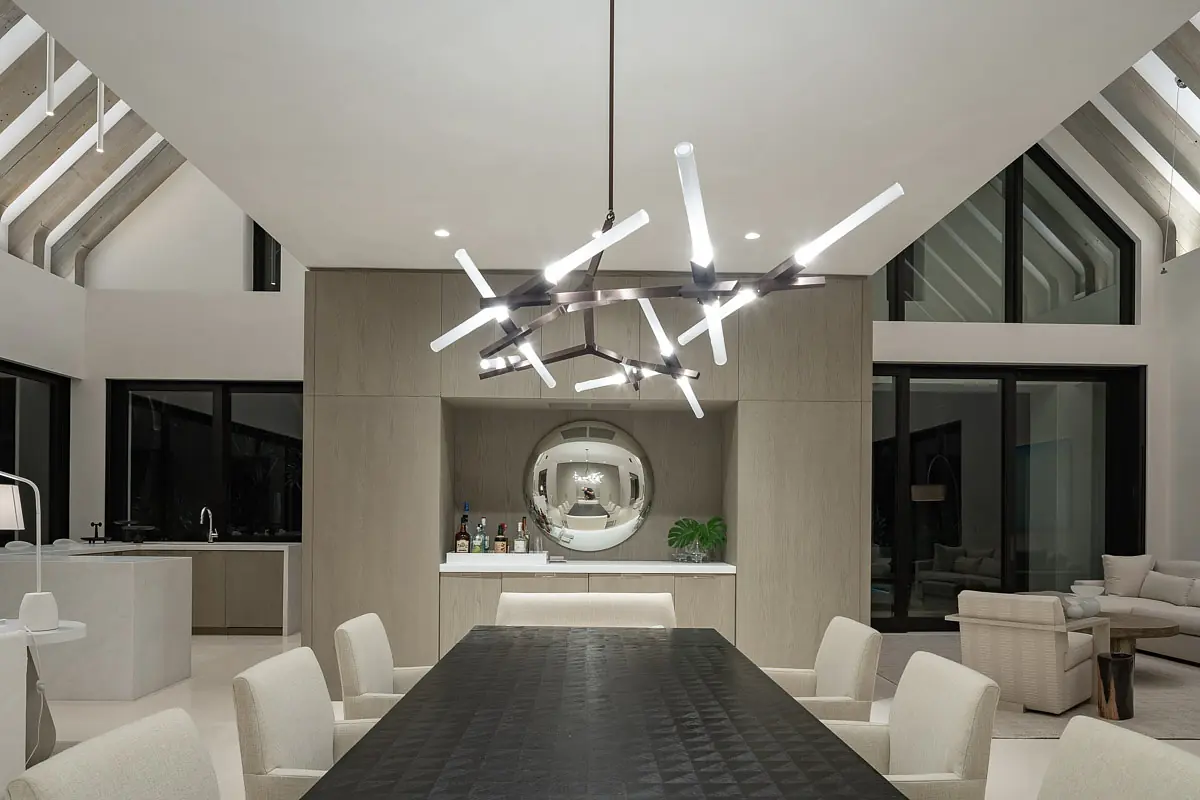 Dining room with large table, beige chairs, built-in bar, round mirror, and floor-to-ceiling windows.