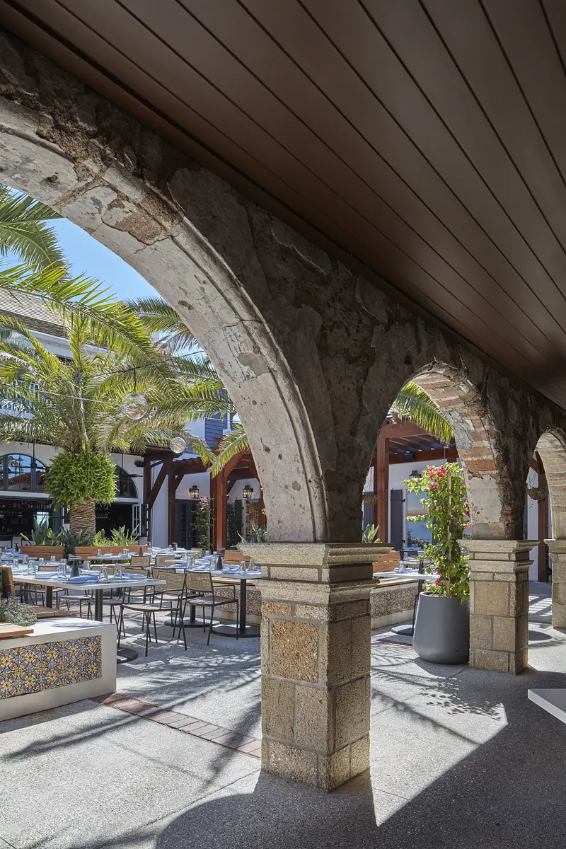 Outdoor patio with stone archways, wooden beams, dining tables, chairs, and potted plants.