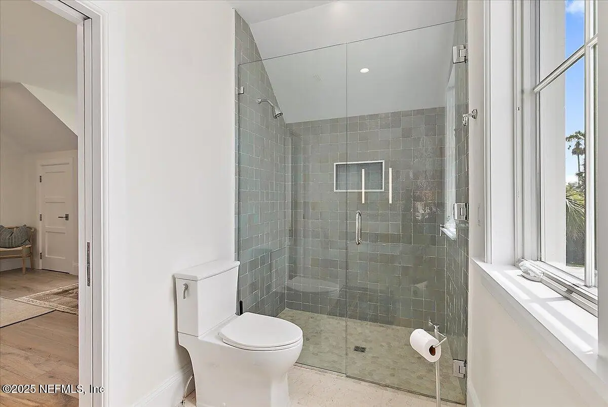 Bathroom with glass shower enclosure, white toilet, gray tiled walls, and natural light from a window