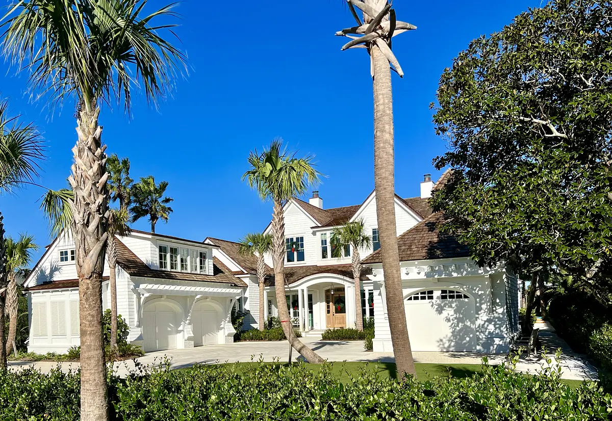 Front exterior view of a house with white siding, brown roof, driveway, palm trees, and landscape.