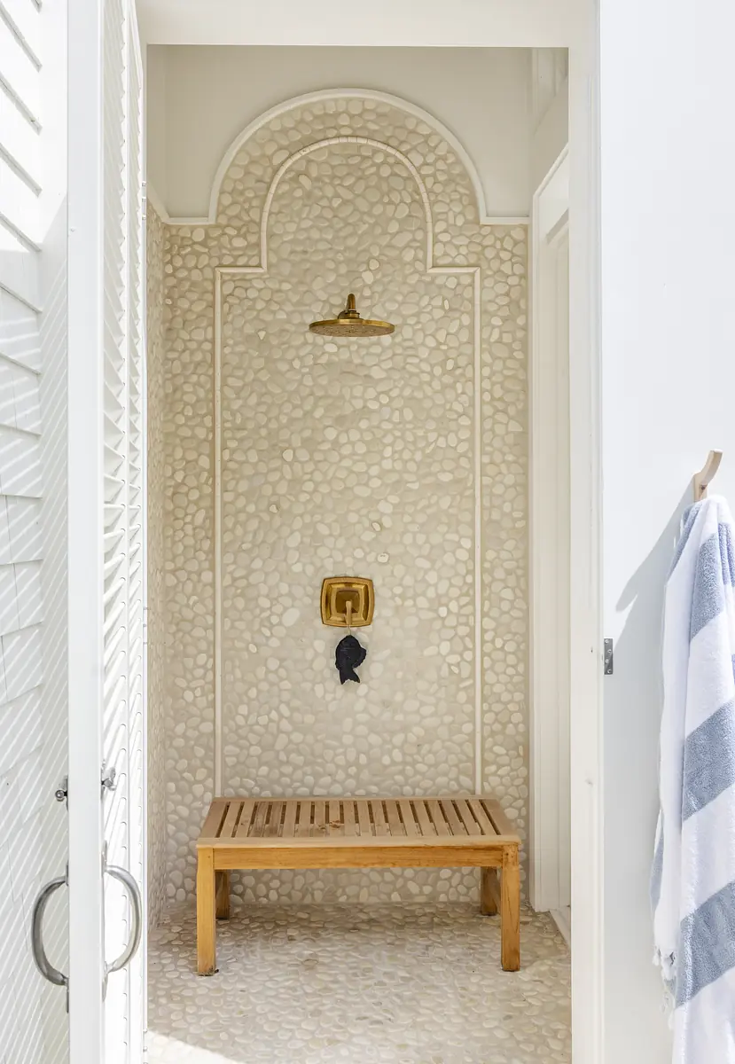 Bathroom with pebble wall, wooden bench, gold shower fixture, and open design.