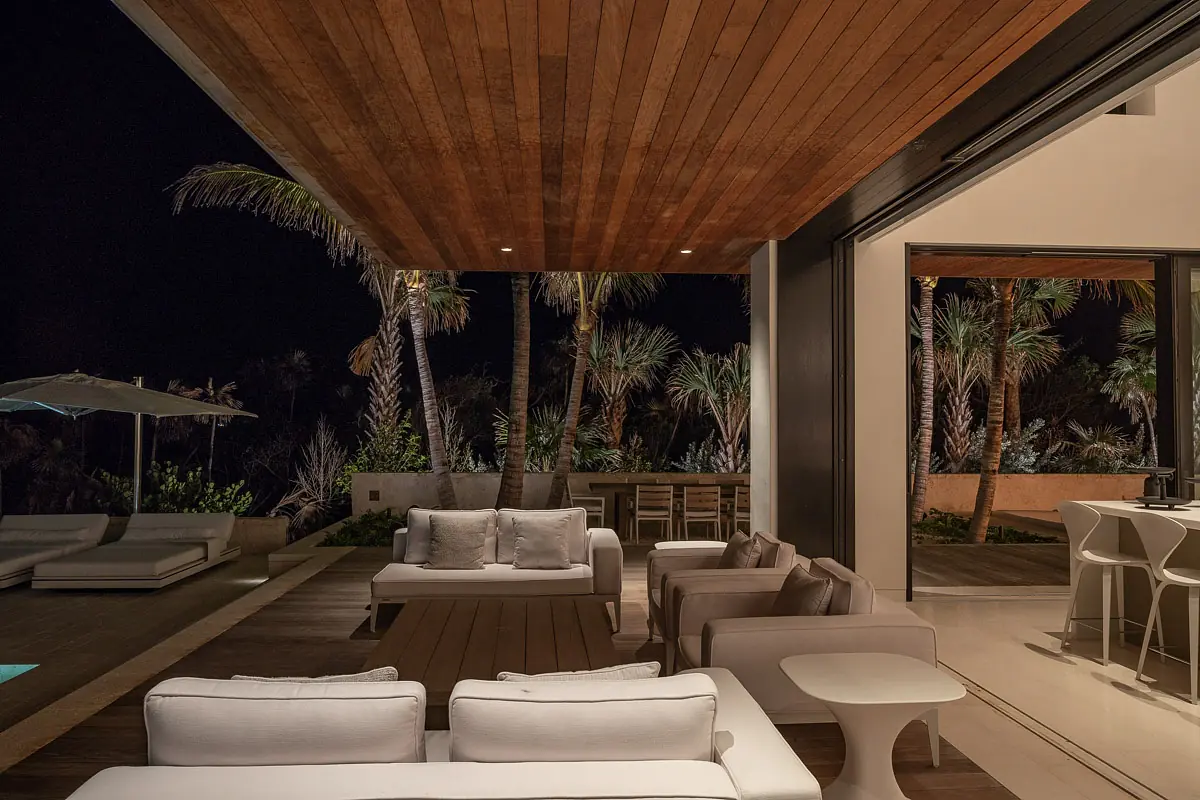 Deck with white sectional sofa, coffee table, bar stools, and outdoor dining table amidst palm trees and a pool