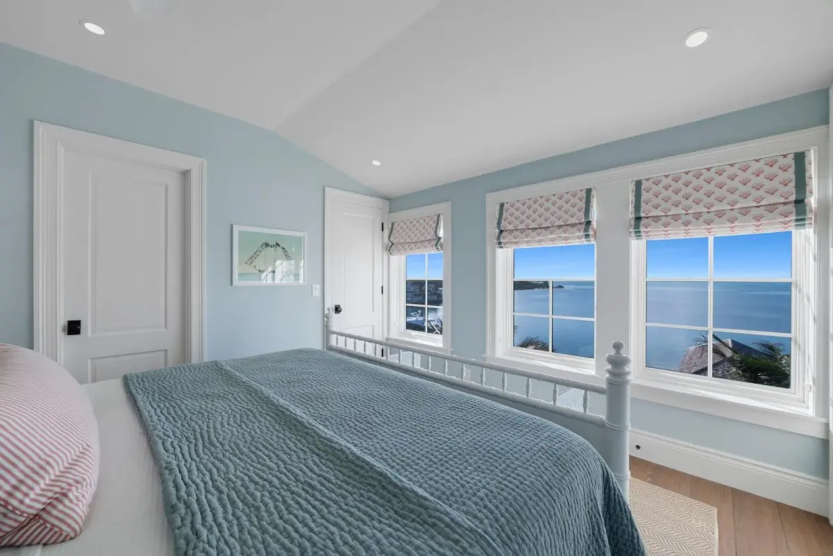 Bedroom with white bed, blue quilt, large windows, and light wood floor.