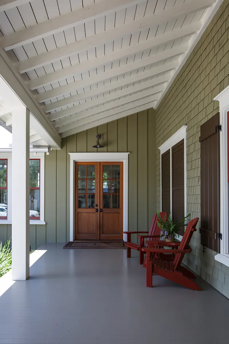 Exterior front porch with gabled roof, red chairs, wooden door, and green shingle walls.