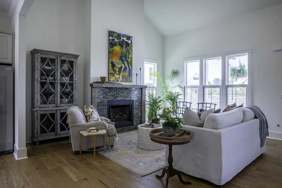 Living room with white sofa, gray armchair, coffee table, fireplace, large windows, and potted plants.