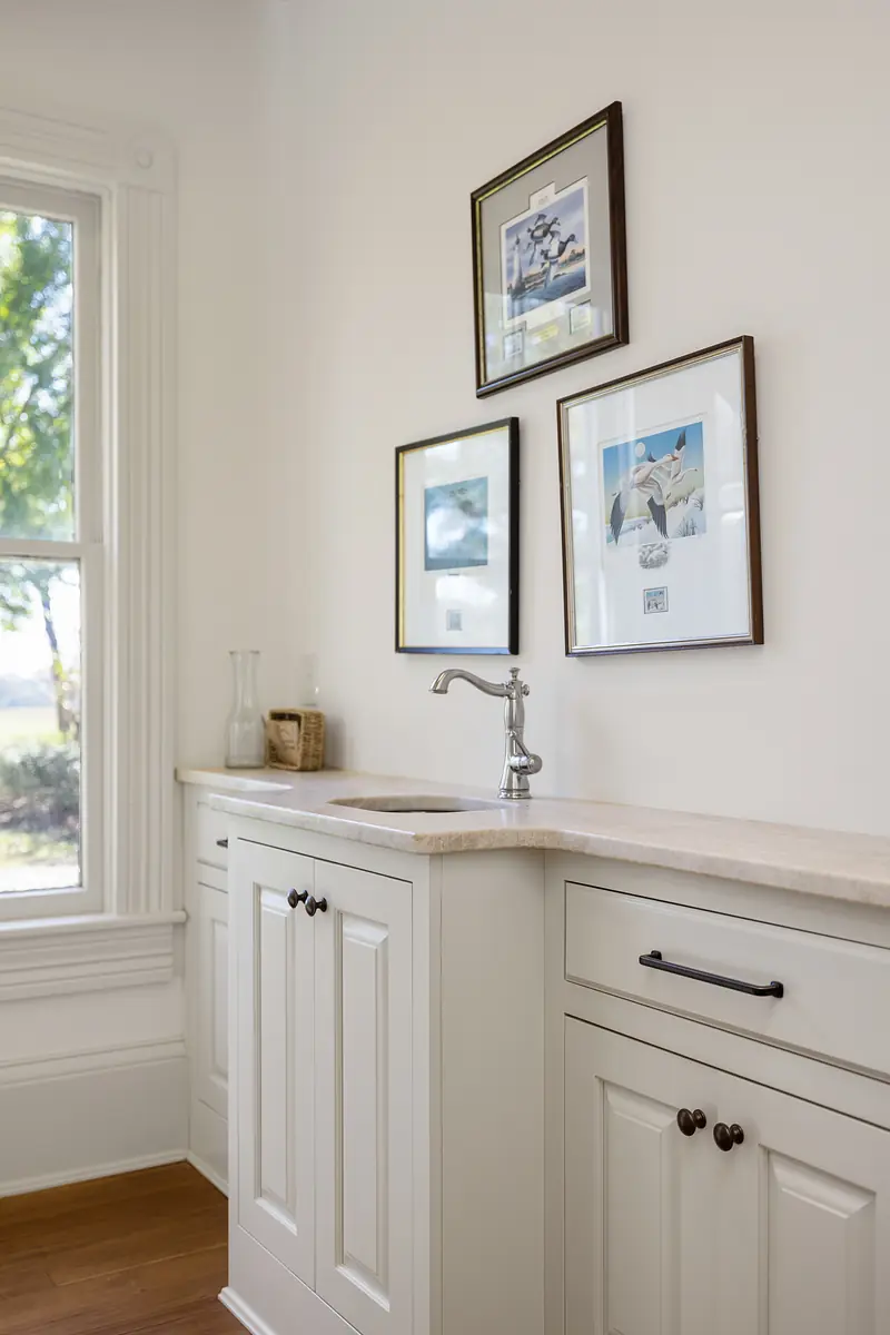 Utility area with light-colored countertop, white cabinetry, sink, and framed artworks near a large window.