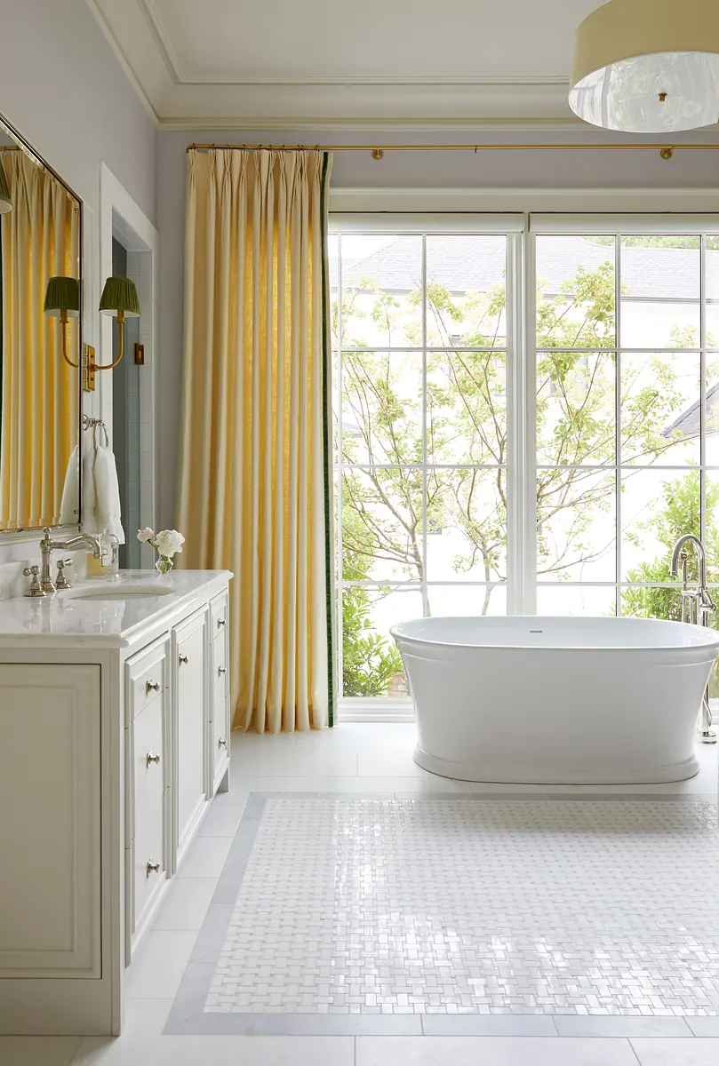 Bathroom with freestanding white bathtub, double vanity, wall mirror, large windows, and yellow drapes.