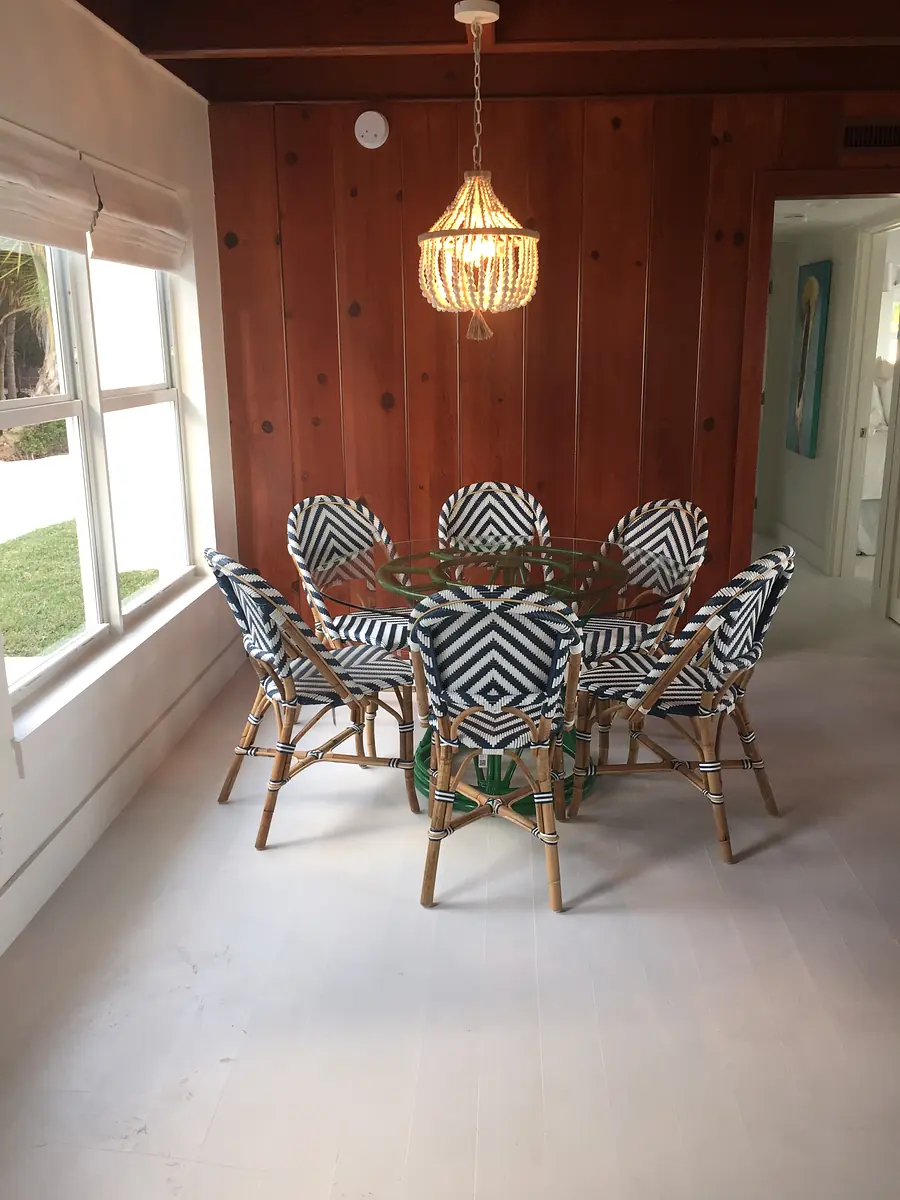 Dining room with round glass table, rattan chairs, wood walls, large window, and pendant light.
