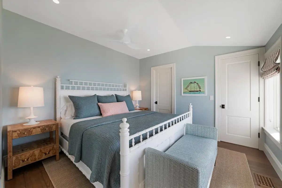 Bedroom with white bed, blue quilt, nightstands, lamps, blue bench, artwork, and natural light from the window