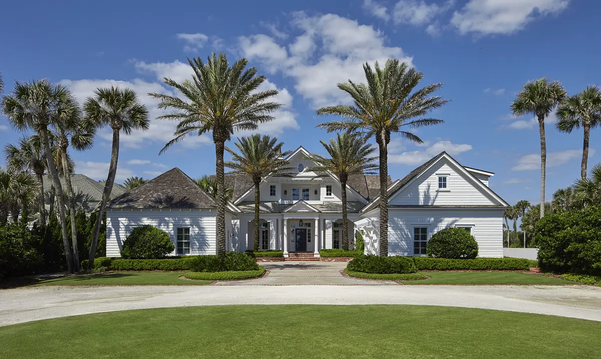 Two-story white house with gabled roofs, palm trees, landscaped greenery, and a central entrance on the front porch.