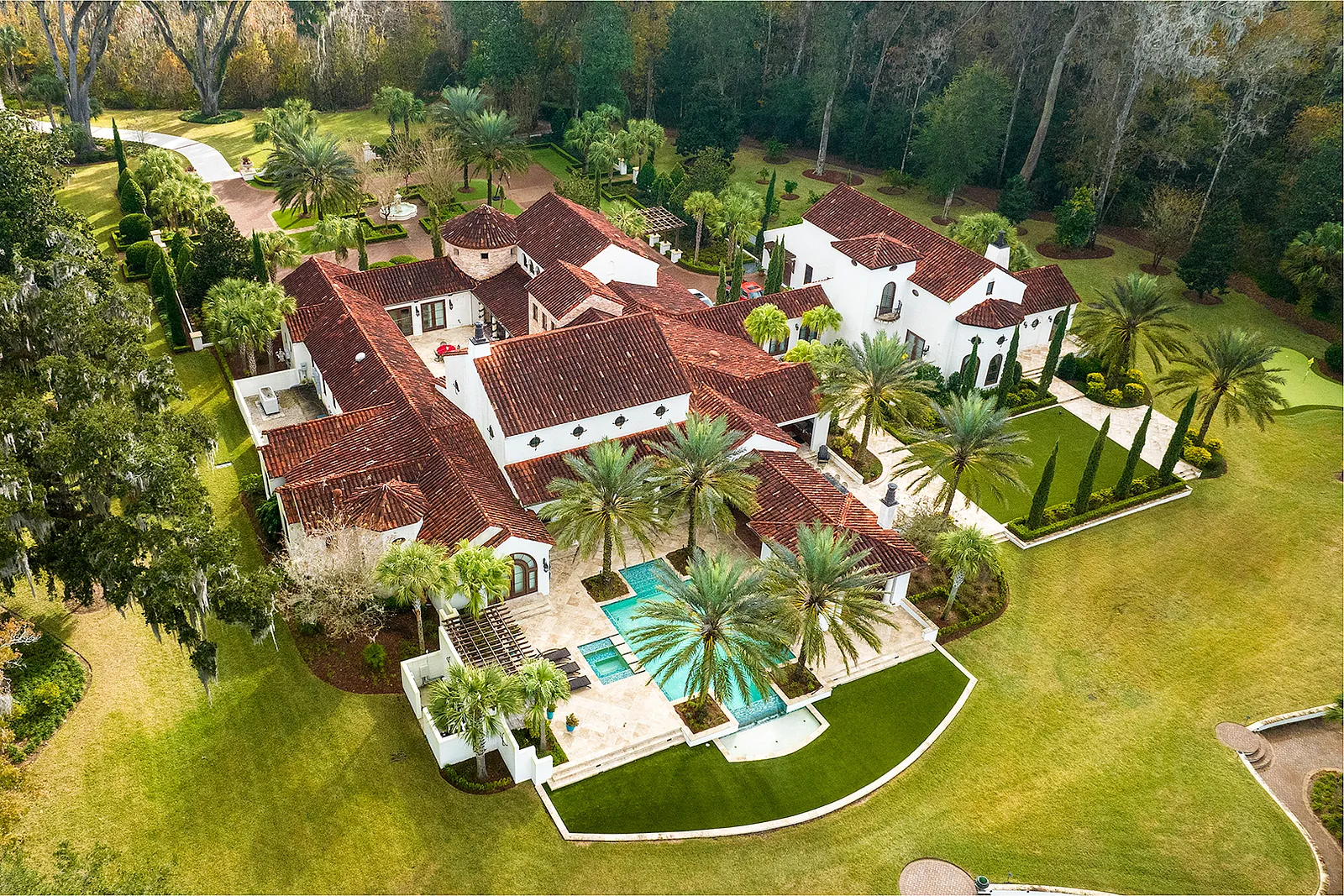 Exterior view of a large estate with terracotta roofs, palm trees, landscaped gardens, and a swimming pool.