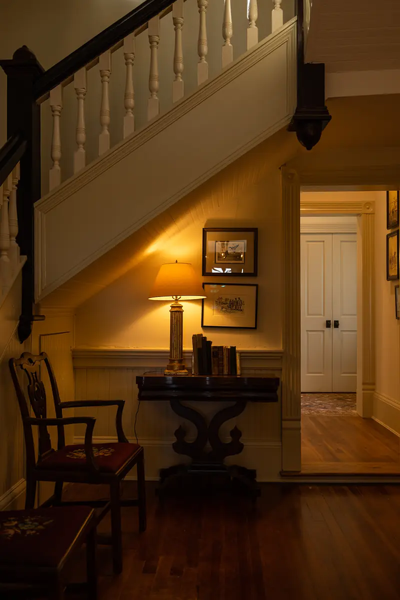 Foyer with wooden table, lamp, books, chairs, staircase, wainscoting, and framed artwork.