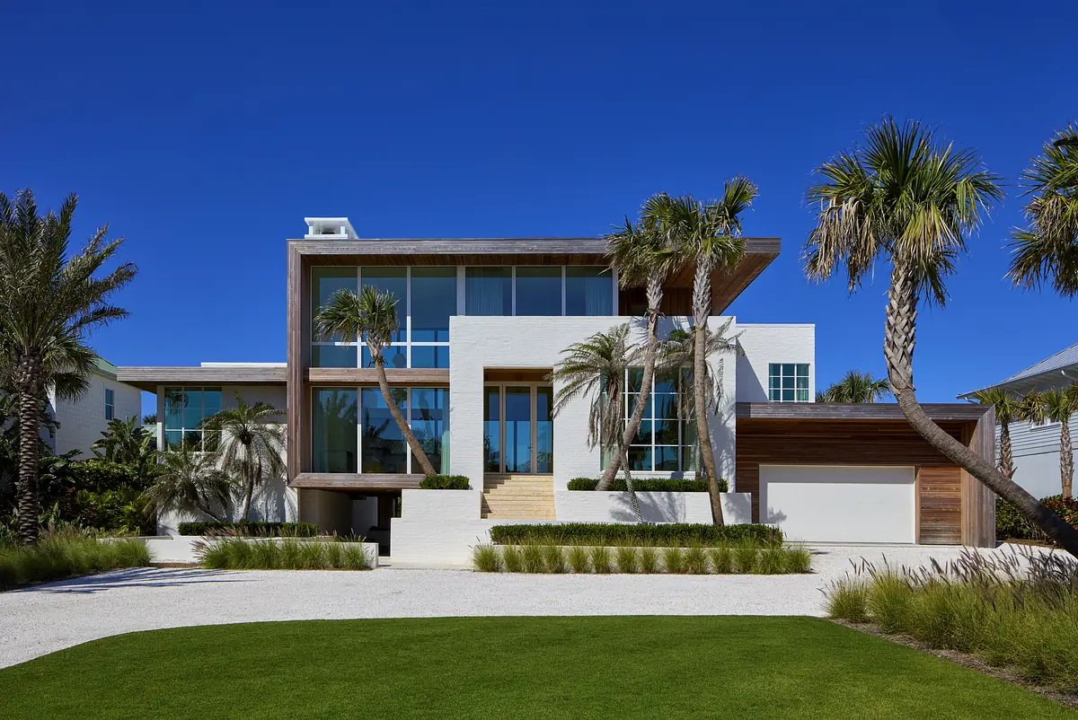 Modern house exterior with white stucco, wood paneling, large windows, and a gravel driveway.