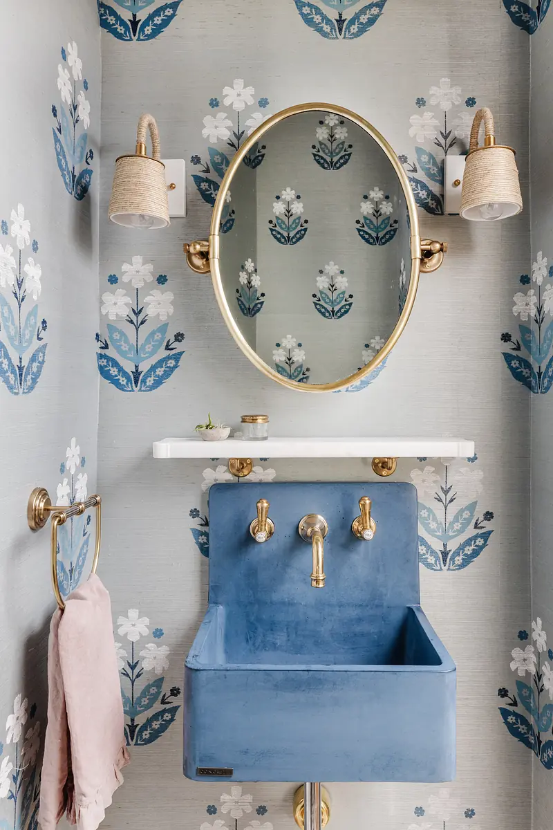 Powder room with blue sink, marble shelf, oval mirror, floral wallpaper, and brass fixtures.