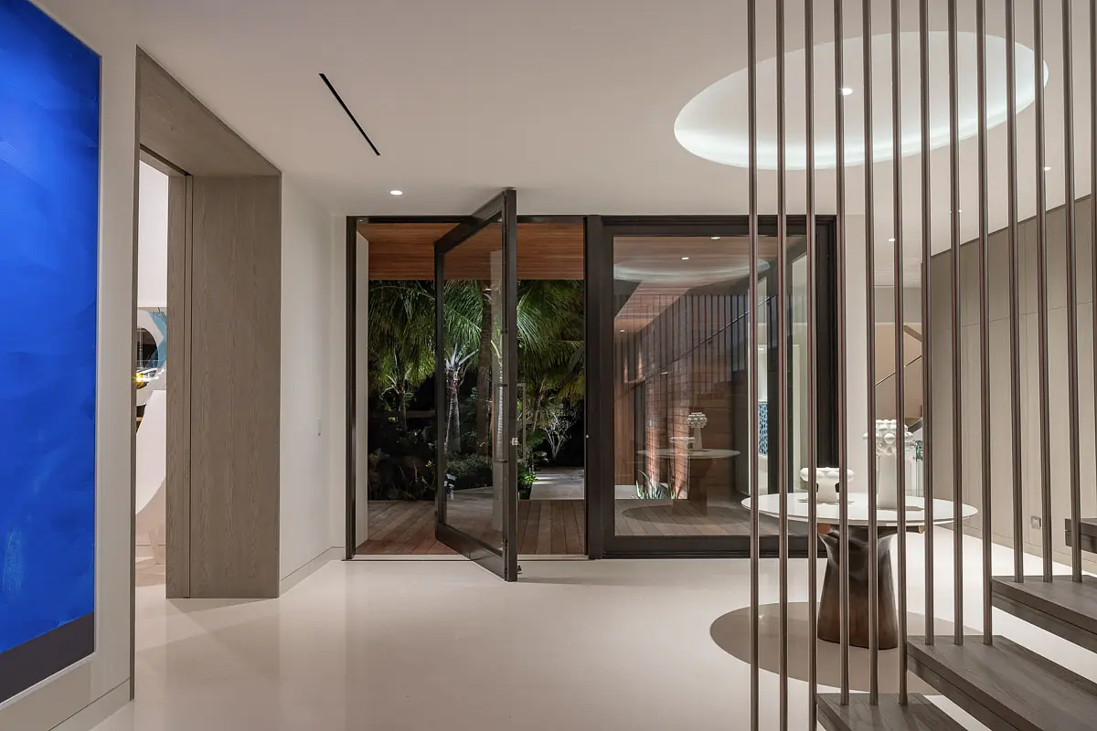 Foyer with glass door, circular table, greenery backdrop, polished floor, modern staircase, and blue artwork.