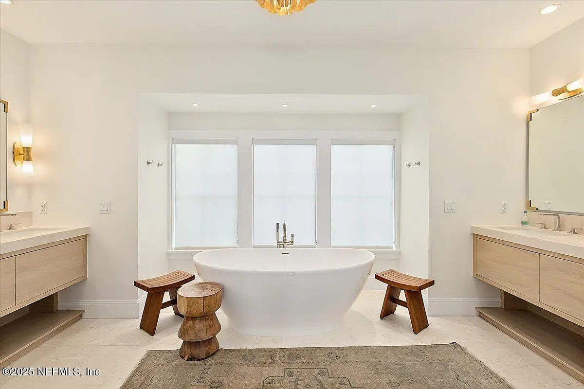 Bathroom with freestanding white bathtub, two vanity sinks, wooden stools, and natural light from windows.