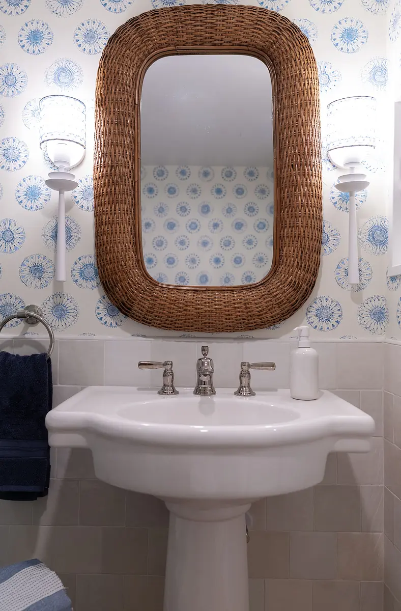 Bathroom with white pedestal sink, woven mirror, blue floral wallpaper, and wall sconces