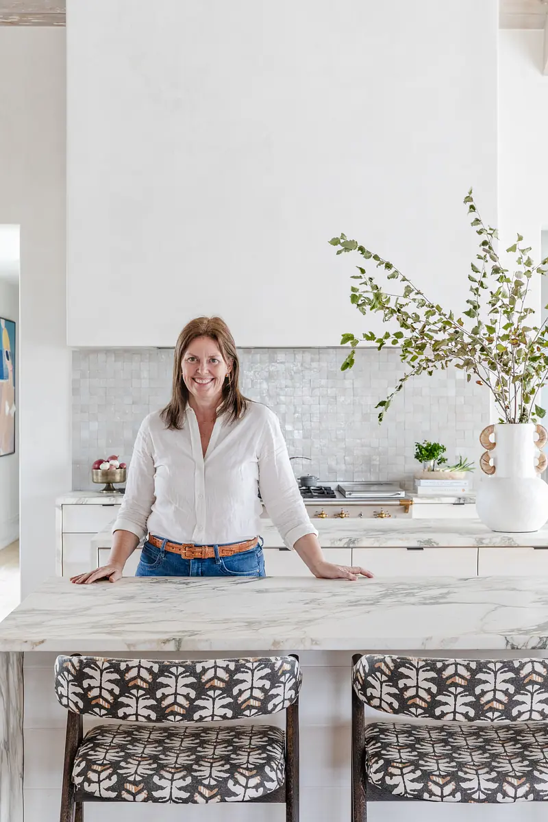 Kitchen with marble countertop island, patterned bar stools, white tiled backsplash, stove, and decorative vase with branches.