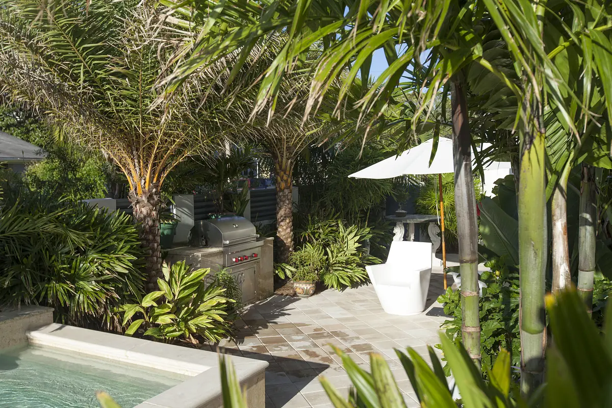 Outdoor patio with white chair, grill, stone table, pool, and tropical plants.