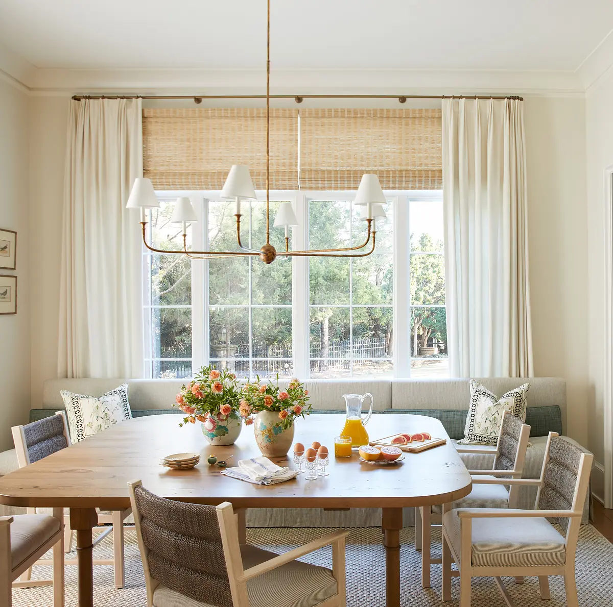 Dining room with wooden table, white chairs, flowers, glassware, bamboo shades, and a light fixture.