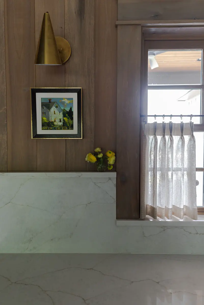 Kitchen area with marble countertop, wood paneling, framed painting, floral vase, and window with sheer curtains.