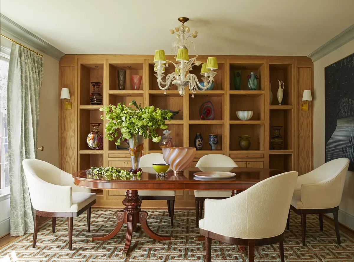 Dining room with round wooden table, white upholstered chairs, shelving unit, and chandelier.