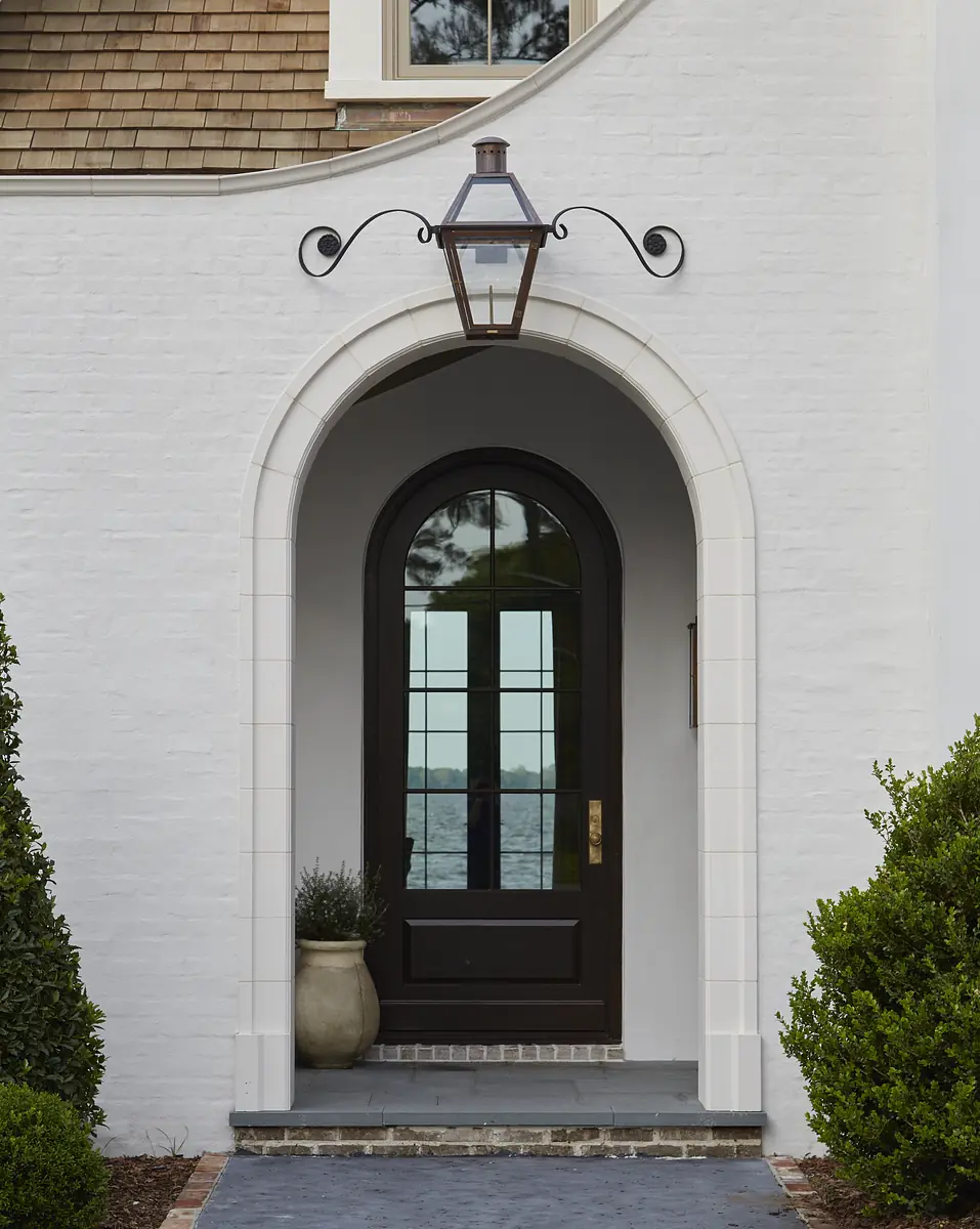 Exterior front with arched doorway, dark wooden door, wall lantern, and flanking shrubs in planters.