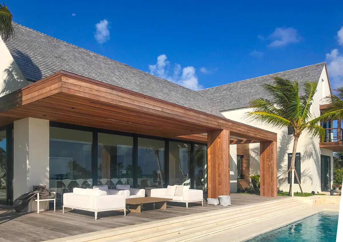 Outdoor patio with wooden overhang, white sofas, coffee table, pool, and palm trees.