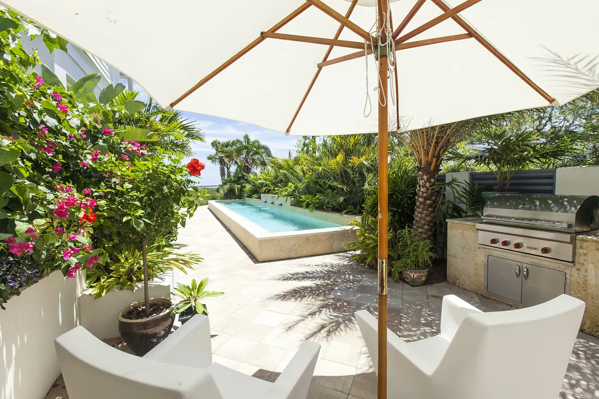 Patio with white chairs, umbrella, swimming pool, grill, and tropical plants.