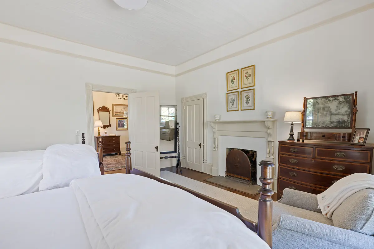 Bedroom with two white beds, wooden dresser, armchair, fireplace, and a door leading to another room.