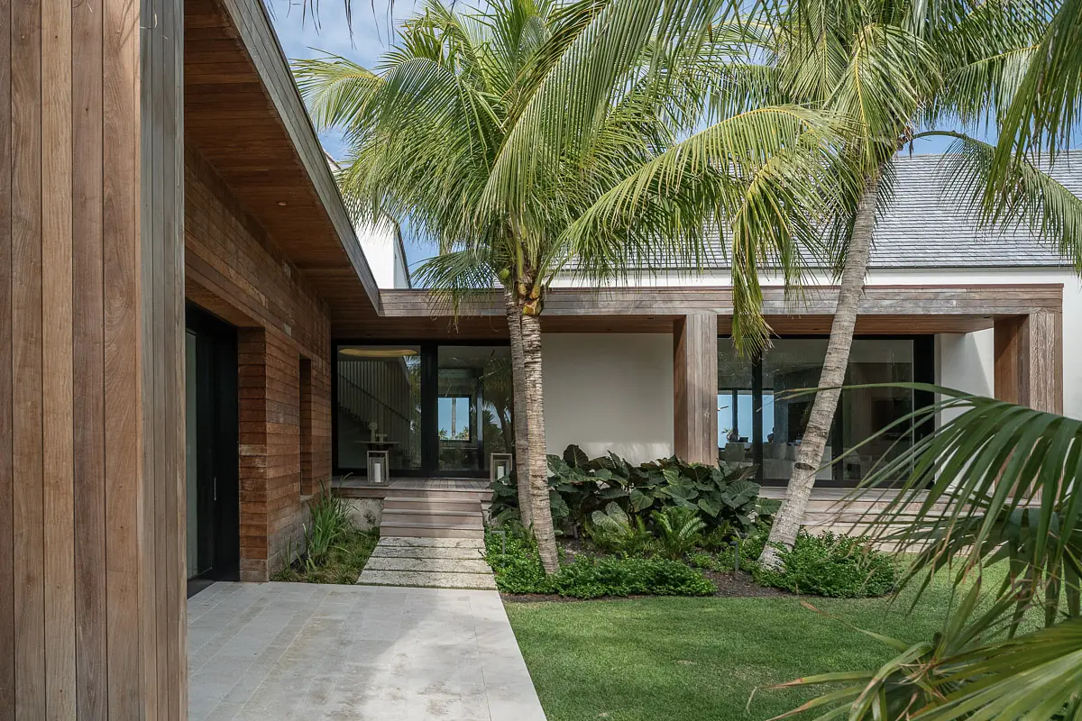 Exterior front view of a modern house with wooden siding, glass windows, palm trees, and stone pathway.