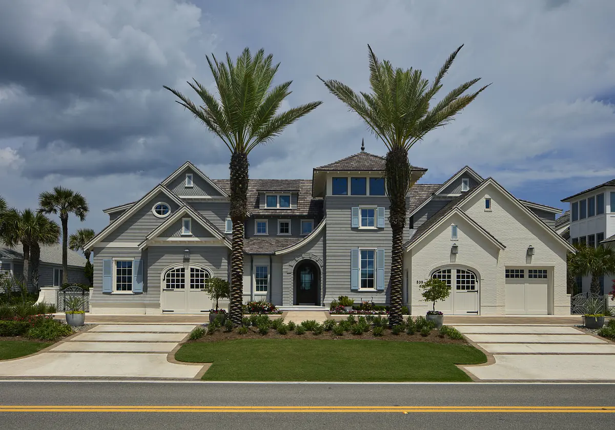 Front exterior of a large house with light gray siding, palm trees, and a manicured lawn.