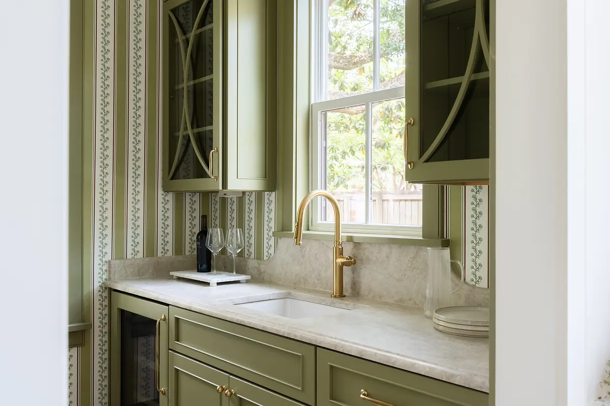 Kitchen area with green cabinetry, marble countertop, gold faucet, wine glasses, and patterned wallpaper.