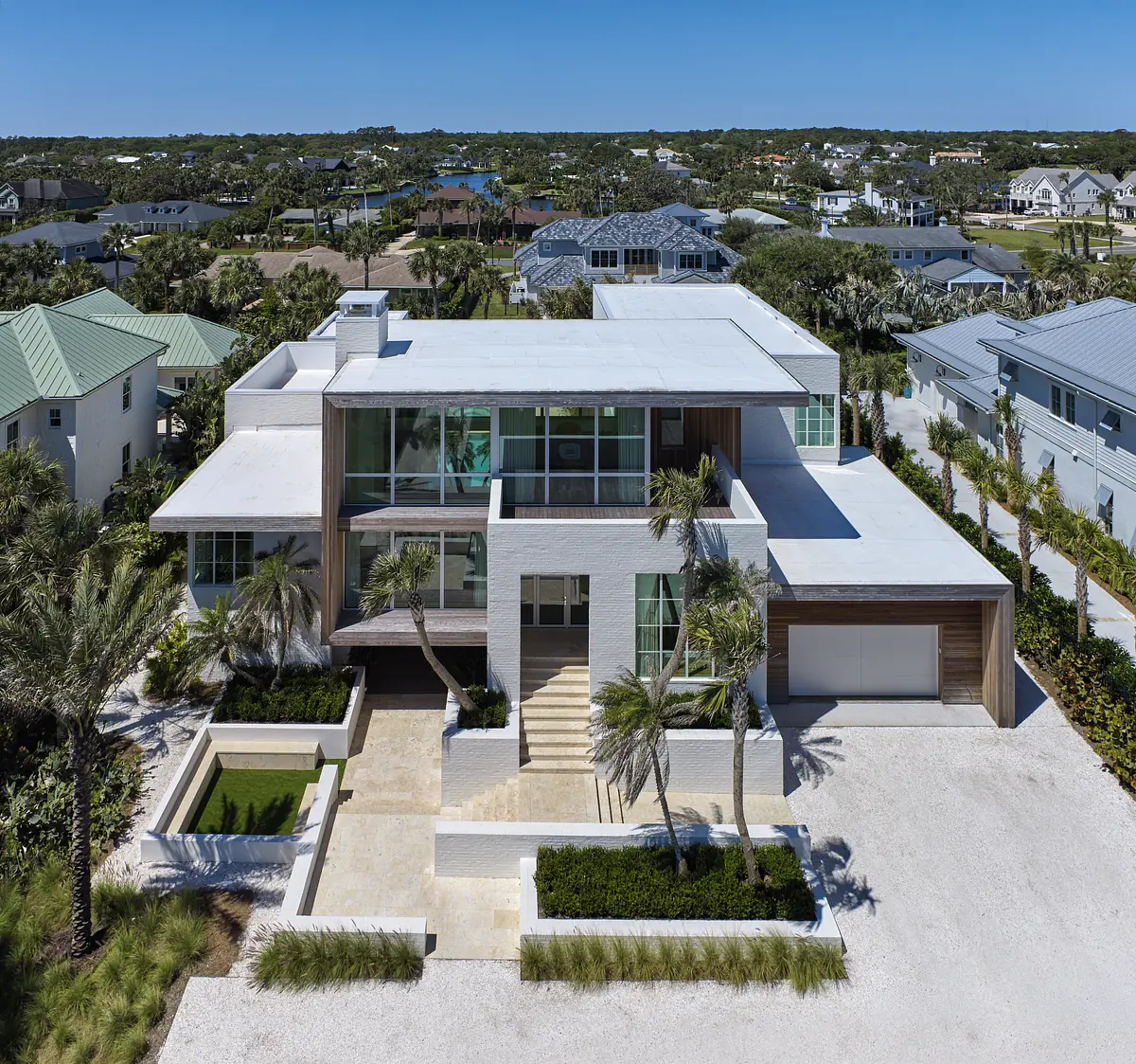 Modern house exterior with large windows, flat roof, palm trees, and landscaped area with a water feature.