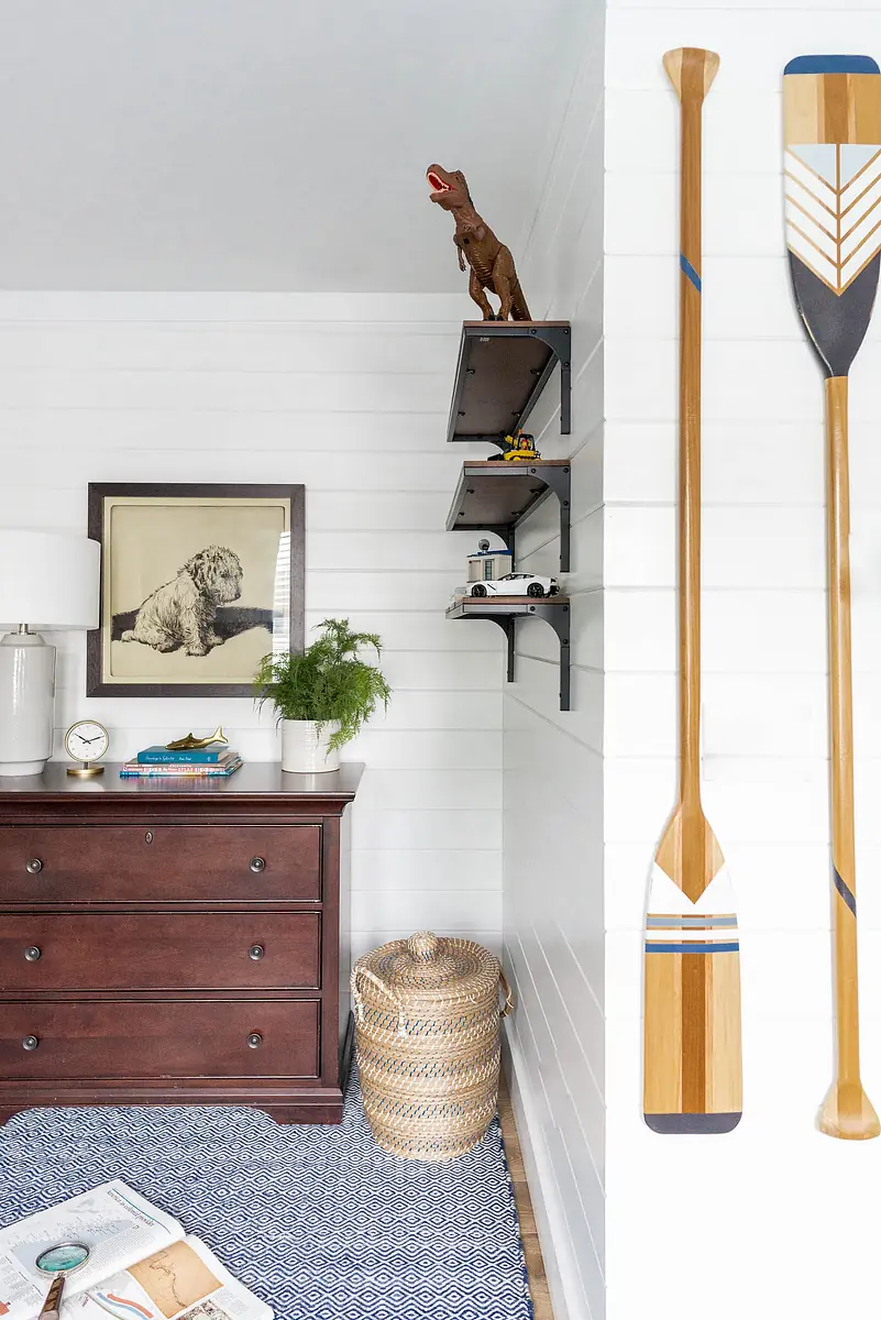 Room with brown dresser, wall shelves, decorative oars, plant, and patterned area rug.