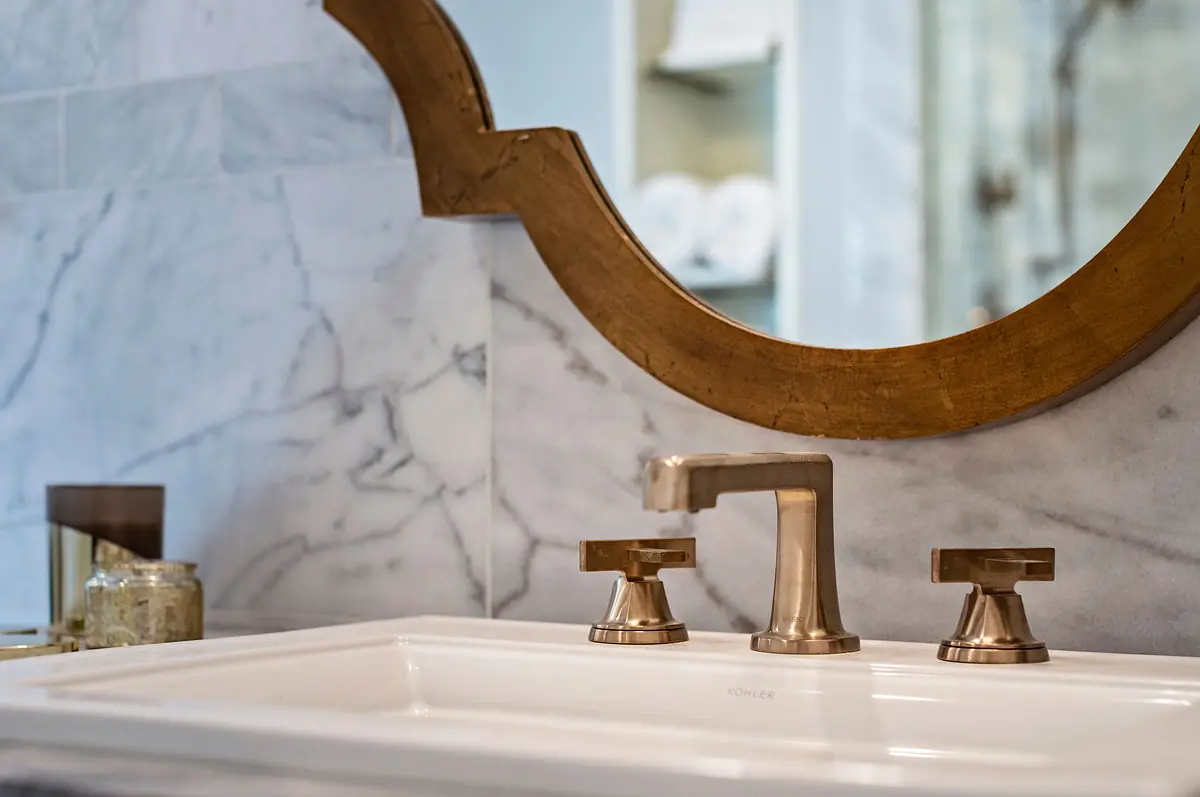 Bathroom with white sink, gold fixtures, round mirror, and marble backsplash.