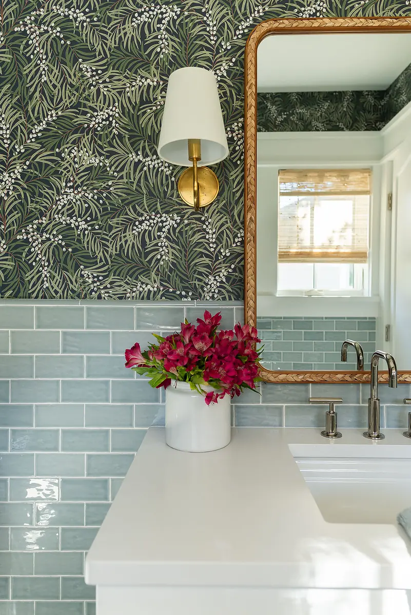 Bathroom with white countertop, blue tile, woven mirror, wall sconce, and vase with pink flowers.