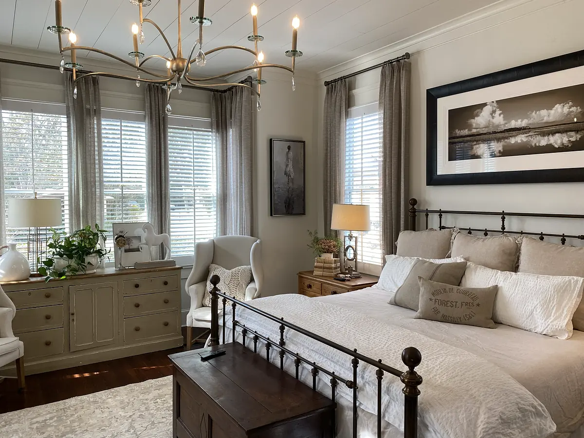 Bedroom with metal framed bed, armchairs, dresser, artwork, and large windows with gray curtains.